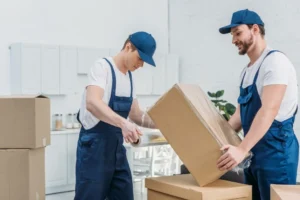 Two professional movers packing and wrapping a large cardboard box in a bright indoor setting, with additional moving boxes nearby